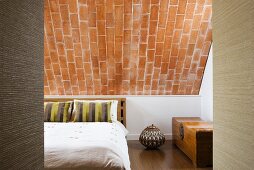Double bed with striped scatter cushions, elegant wooden chest and ceramic pot with geometric pattern under a sloping brick ceiling