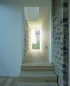 Wooden stairs next to a stone wall leading to a narrow room with shelving opening to a gallery above and with a glass door at the end