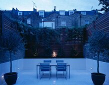 Dusk in a small, English courtyard with spotlights recessed in white wall cladding contrasting with high wooden fence with climbing plants
