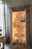 Crockery in illuminated, glass-fronted cabinet with open doors against grey-painted wall