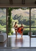 High-ceilinged room with view of landscape through open glass wall - two girls in bikinis with towels on way to pool