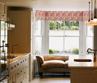 Cream-coloured, retro-style country house kitchen with leather couch in bay window and floral Roman blind