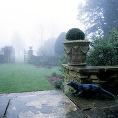 Box ball in vintage planter and stone fox statue on terrace with view into misty garden
