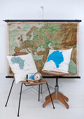 Cushions with cartographic motifs on chair and wooden swivel chair in front of map hanging on wall