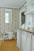 Country-style kitchen with patterned wallpaper and white crockery on kitchen counter with curtained front below wall brackets with lace trim