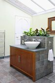 Modern bathroom with glass roof and grey tiles; wooden doors on central washstand island with integrated planter