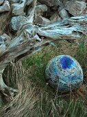 Blue-glazed ceramic sphere with ribbon of text lying in grass next to gnarled root wood