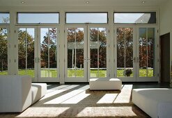 weiße Polsterhocker im minimalistischen Wohnraum und Blick durch Terrassenglastüren auf Garten