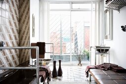 View of city roofscape through floor-to-ceiling glass wall in unconventional bathroom with stainless steel frames and leather couch