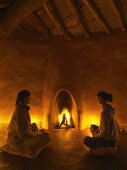 Meditative atmosphere in simple clay building with organic lines - two women sitting on floor cushions in front of blazing fire in fireplace
