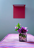 Glas mit Bougainvillea-Blüten auf gestreiftem Polster in Rot/Violett; im Hintergrund rotes Kunstobjekt vor taubenblauer Wand