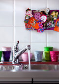 Colourful plastic beakers next to stainless steel sink with running water below souvenirs on pinboard