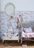 Soft toys in old doll's cradle next to vintage wicker chair against wall with half-height Toile de Jouy wallpaper