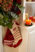 Gestrickte Nikolaussocken unter Blumendeko mit Rosen und Hortensien, daneben brennende Kerze auf Wohnzimmerschrank