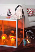 Lit candles and Christmas baubles in large floor lantern with orange glass next to sofa with pale upholstery