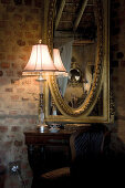 Table lamp on antique console table in front of gilt-framed mirror on brick wall