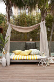 Daybed with fabric canopy hung between palm trees on modern wooden terrace