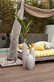Collection of vases on floor of wooden terrace next to modern daybed with fabric canopy hung between palm trees