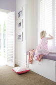 Girl sitting on window seat built into niche of window with closed, white, slatted shutters