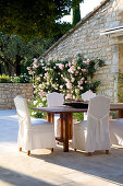 Chairs with loose covers and wooden table on terrace with climbing rose on stone facade of French country house