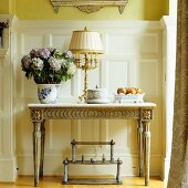 Plant pot and table lamp on antique console table with gilt carved ornamentation against white wood panelling