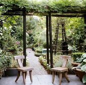 Rustikal moderne Holzstühle auf Terrasse unter berankter Pergola und Blick in blühenden Garten