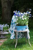 Blaue und weiße Sommerblumen in alter Emaille-Schüssel auf Bistrostuhl und gemusterte Tischdecke über der Rückenlehne