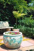 Seerosenbecken in Tontopf mit bunter Mosaikverkleidung auf gepflastertem Sitzplatz im Garten