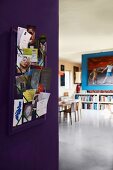 Photos and cards on pinboard on purple corridor wall with view into modern dining room beyond