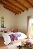 Sichtbare Dachkonstruktion mit Strohmatten in freundlich hellem Schlafzimmer; Kissen und Decken in Rotlilatönen auf französischem Bett