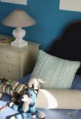 Soft toys, bolster and cushion on bed next to lamp on chest of drawers