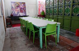 Long wooden table with peeling paint and green plastic chairs in front of wooden partition with stained glass windows in simple dining room