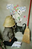 Child with straw basket on head sitting on floor next to besom leaning on wall with children's motifs