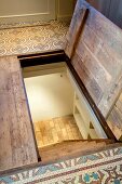 Halboffene Kellerluke in Fussboden mit gemusterten Fliesen und Blick auf steile Treppe