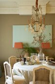 Dining table and antique chairs below crystal chandelier; delicate colour scheme with salmon pink lampshades against pastel green and light brown