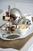 Biscuits and packaged chocolate bars on a tiered cake stand next to assorted sugar bowls on a vintage porcelain platter