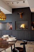 Elegant designer dining area in front of dark wall of fitted cupboards with integrated fireplace below deer-head sculpture