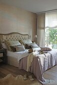 Clothing on double bed with curved headboard next to large window with view of garden