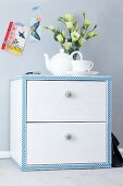 Small chest of drawers decorated with patterned tape with vase of Texas bluebells and romantic postcards on wall