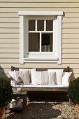 Greyed, weathered teak bench with white cushions below window of wooden house