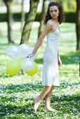 Young woman walking in field of flowers, holding balloons behind back, looking over shoulder