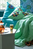 Tea set on side table in front of bed with colourful cushions and bedspread against wall with Oriental patterned tiles