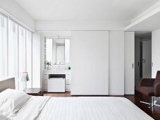 Doppelbett vor der Fensterfront in weißem Schlafzimmer mit Bad ensuite hinter Schiebetüren