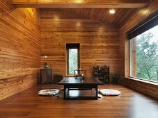 Asiatisches Teezimmer mit Bodenkissen am niedrigem Tisch aus dunklem Holz im holzverkleideten Wohnraum mit grossem Fenster