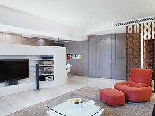 Moderner Wohnraum mit rosa Polstersessel und passendem Fussschemel gegenüber Raumteiler mit integriertem Fernseher