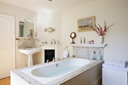 Bathtub accessible from three sides clad in white-painted wood and pedestal sink next to small fireplace