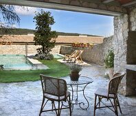 Basketwork chairs and metal bistro table on enclosed stone terrace with loungers and pool in background