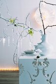 Artistic Christmas atmosphere - glass icicles hanging from twigs in white china vase on cubic side table against wall lit with fairy lights