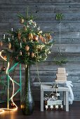 Decorated Christmas tree in vase next to presents on stool against simple wooden wall