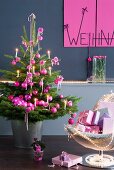 Geschmückter Weihnachtsbaum mit pinkfarbenen Kugeln neben Sessel mit Geschenken vor grauer Wand und Weihnachtsgruss an pinkfabener Tafel